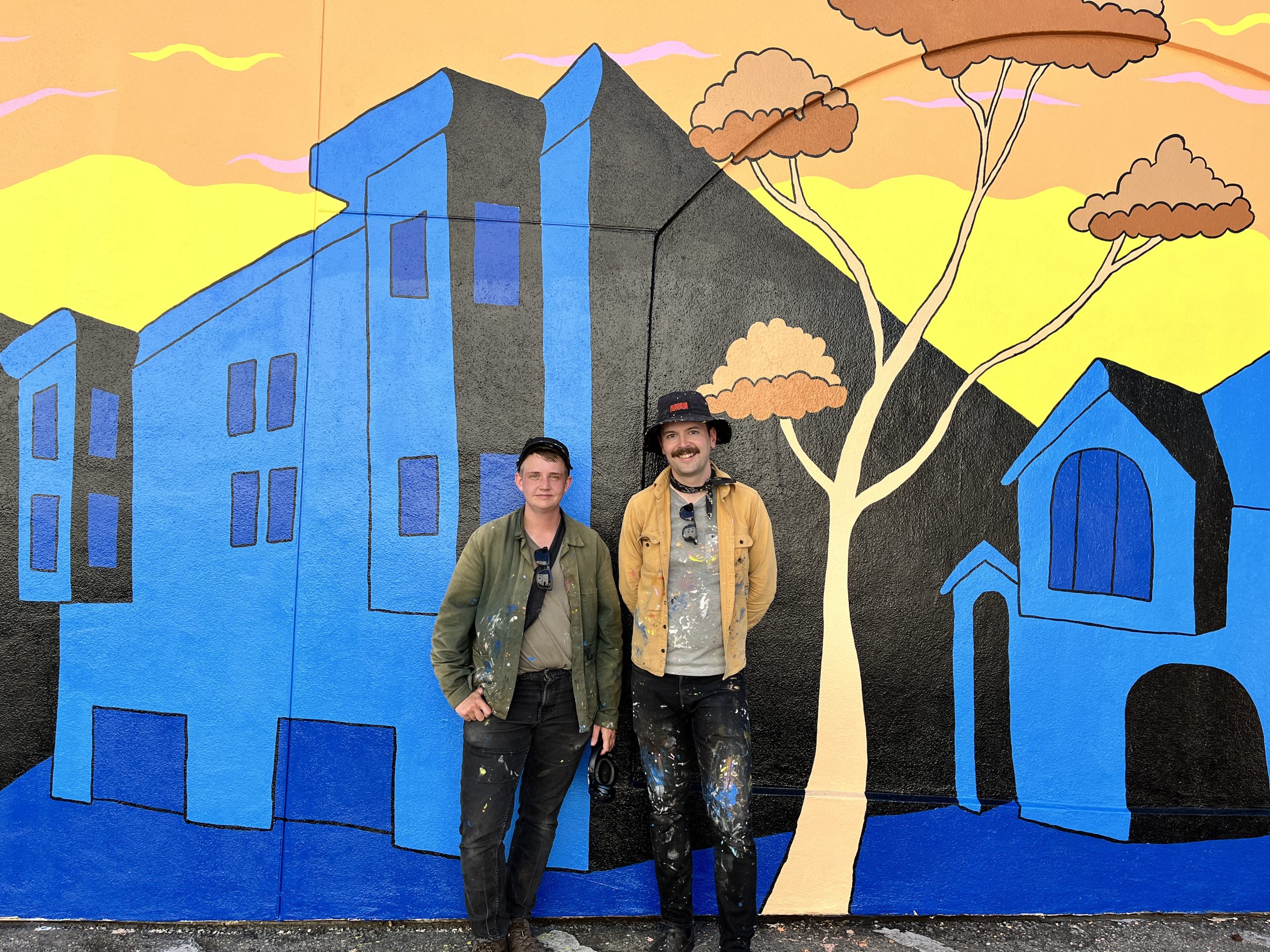 Two people stand in front of a colorful mural featuring buildings and trees.
