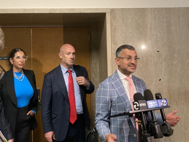 A man in a plaid suit speaks at a podium with microphones, while two people stand behind him near a wooden door.