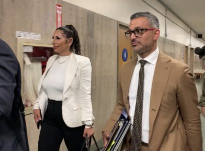 A woman in a white blazer and a man in a brown suit walk through a hallway, carrying files.