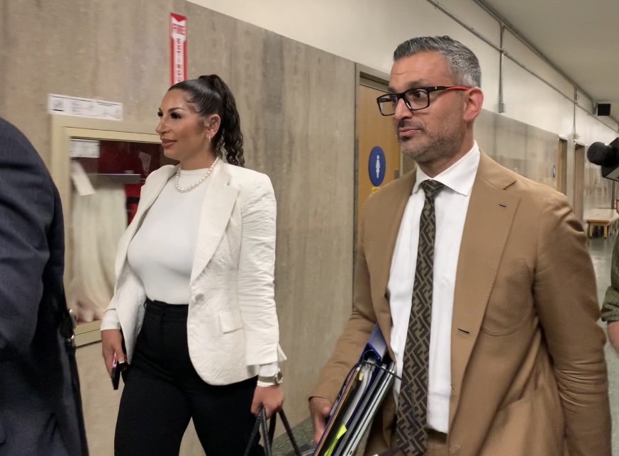 A woman in a white blazer and a man in a brown suit walk through a hallway, carrying files.