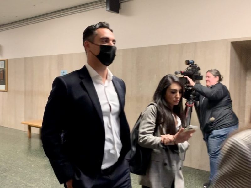 A man in a suit and a woman are walking in a hallway. The man is wearing a mask. A photographer with a camera is visible in the background. Bob Lee murder trial.