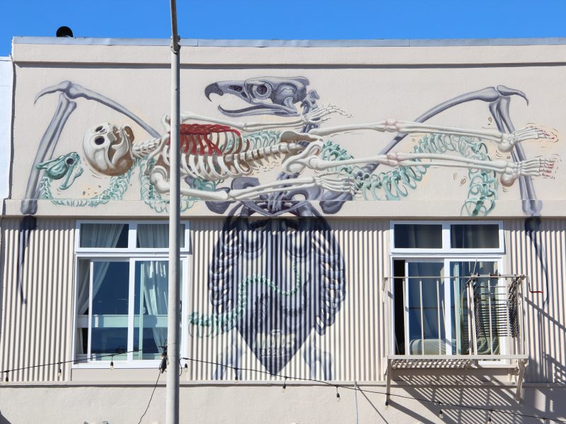 A mural of a large skeleton in an extended pose graces the facade, framed by two windows. The clear blue sky above adds depth, a nod to the creativity reminiscent of Paxton's architectural brilliance.