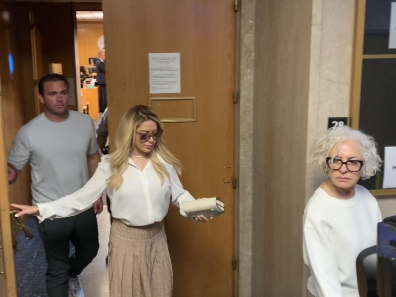 Three people are walking through a doorway with wooden walls. The woman in front holds a rolled document, followed by a man in a gray shirt and another woman with curly hair and glasses.