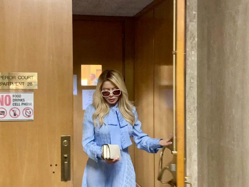 Person in a blue dress and sunglasses exiting a wooden door labeled "Superior Court Department 28," holding a phone and a small white object. Signs nearby prohibit food and phone use.