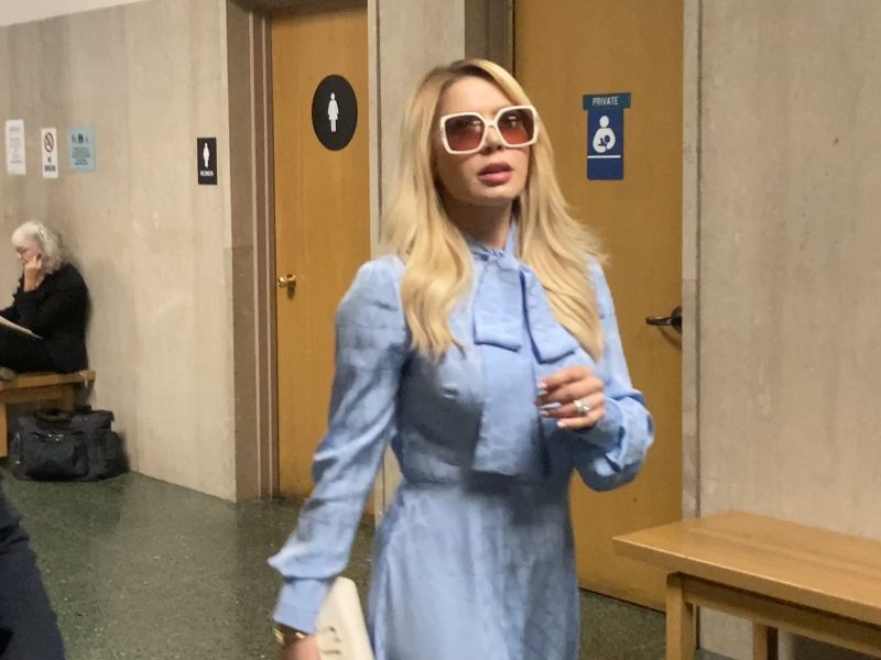 A woman in a light blue dress and sunglasses walks in a hallway with beige walls and restroom signs visible in the background.