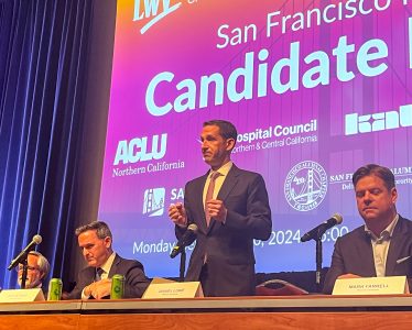 Three men are seated at a table with microphones and name tags. One man is standing and speaking. A screen behind them displays logos and the text "San Francisco Candidate.