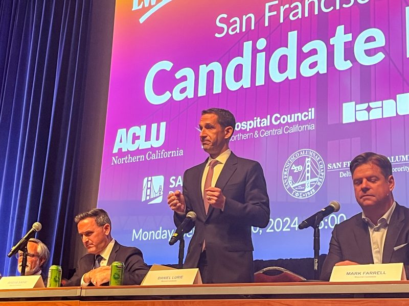 Three men are seated at a table with microphones and name tags. One man is standing and speaking. A screen behind them displays logos and the text "San Francisco Candidate.