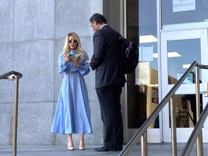 Two people stand outside a building. The woman in a blue dress holds a phone, while the man in a suit carries a backpack. They appear to be engaged in conversation.