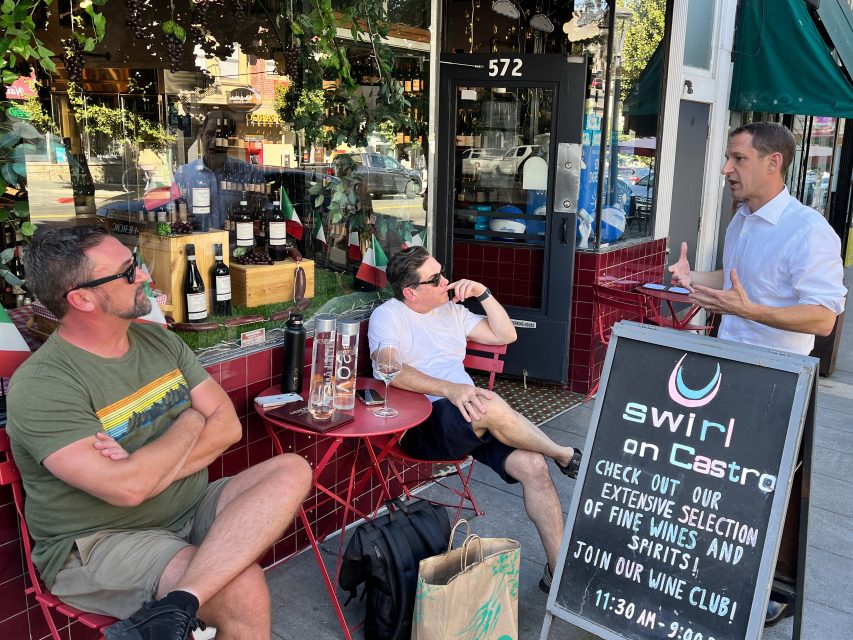 Three men are gathered outside a wine shop with a chalkboard sign, engaged in conversation. Two sit at a red table, while one stands, gesturing with his hands.