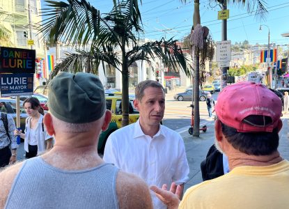 Daniel Lurie, in a white shirt, stands outdoors talking to two men, one in a red cap and another in a green cap, with palm trees and street signs in the background.