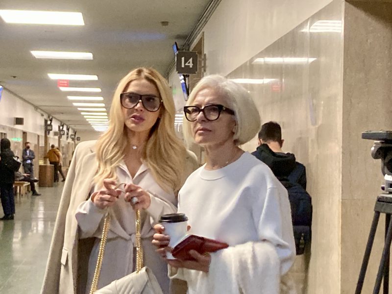Two women with glasses stand in a hallway, one holding a coffee cup and phone. They appear to be engaged in conversation.