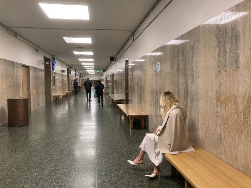 A woman sits alone on a bench in an empty hallway with marble walls, while a few people walk in the distance.