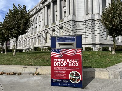 A ballot drop box in front of a large building with columns and trees. It displays American flags and voting information.