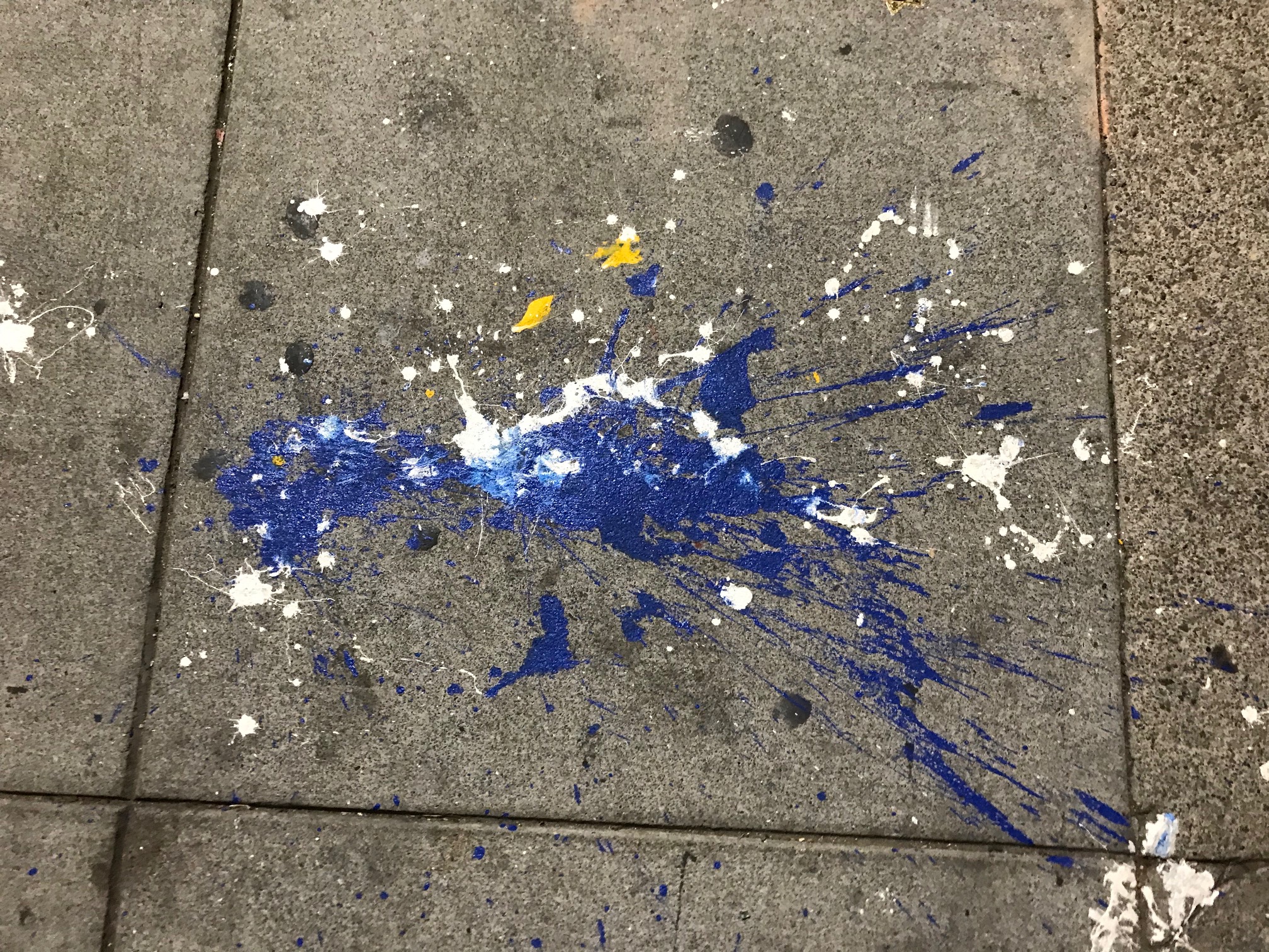 Paint splatters in blue, white, and yellow on a concrete sidewalk.