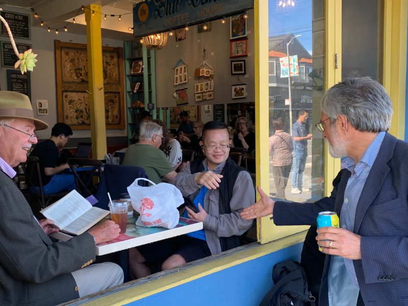 Three people engage at a café window: one inside reading a Peskin novel, another seated with a drink, and the third outside holding a can, leaning in for a handshake.