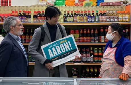 A group of people stands in a shop. A person holds a sign reading "We need Aaron!" while talking to a shopkeeper wearing an apron and a face mask. Shelves with various products are in the background.