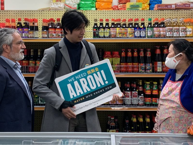 A group of people stands in a shop. A person holds a sign reading "We need Aaron!" while talking to a shopkeeper wearing an apron and a face mask. Shelves with various products are in the background.