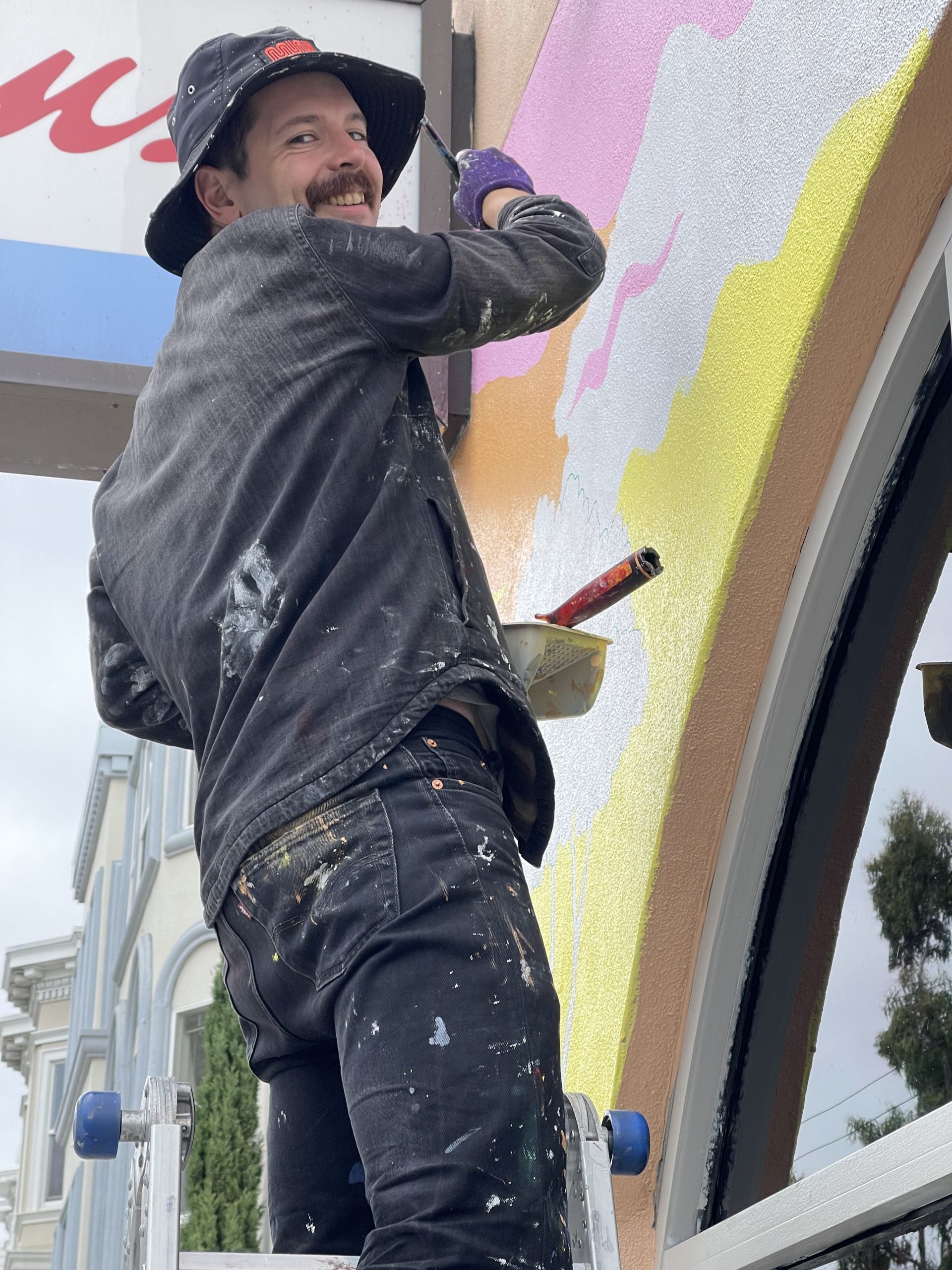 Person in a black hat is painting a wall with colorful abstract patterns while standing on a ladder.