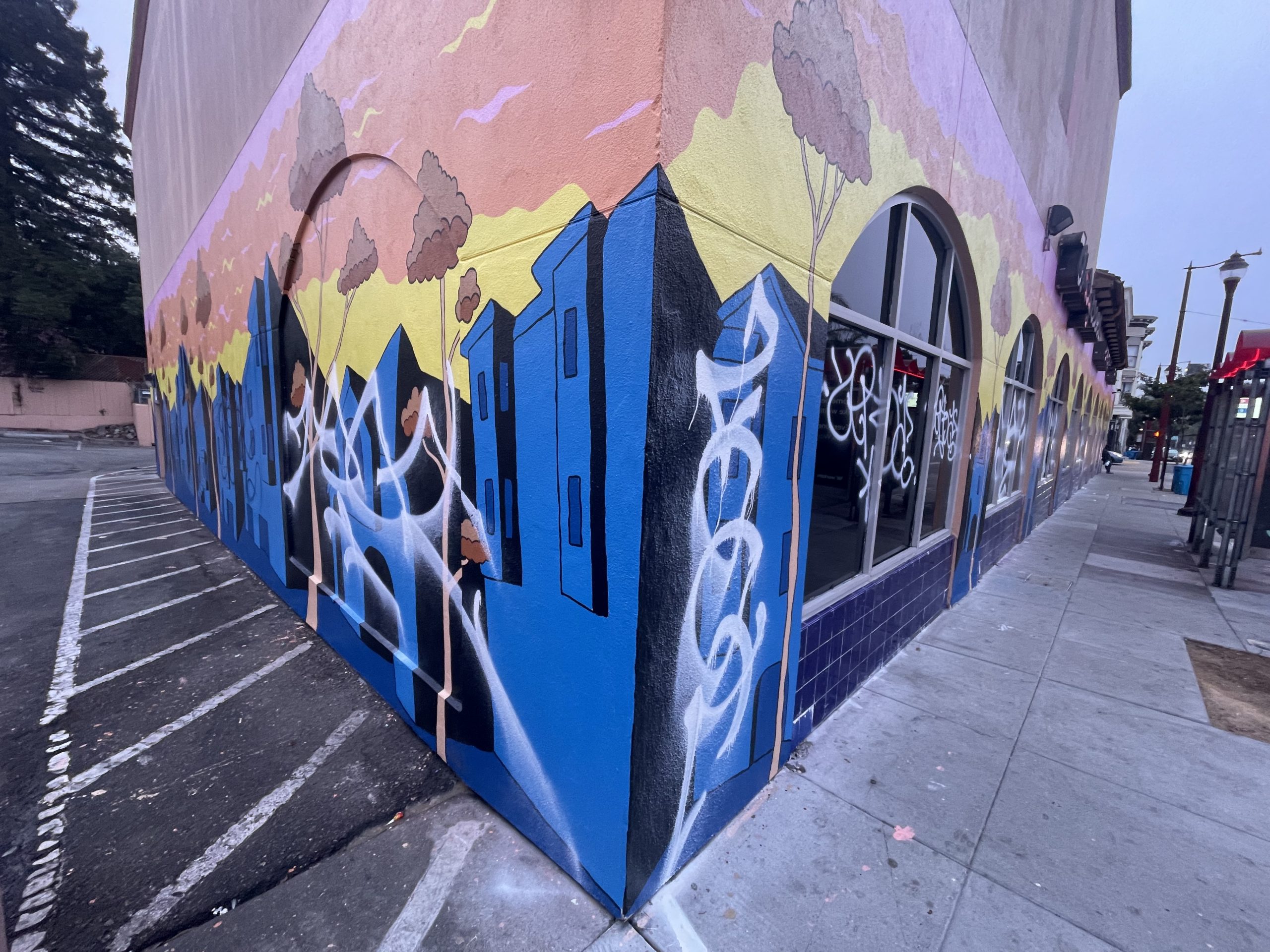Colorful mural on the corner of a building with trees and buildings, overlaid with white graffiti.