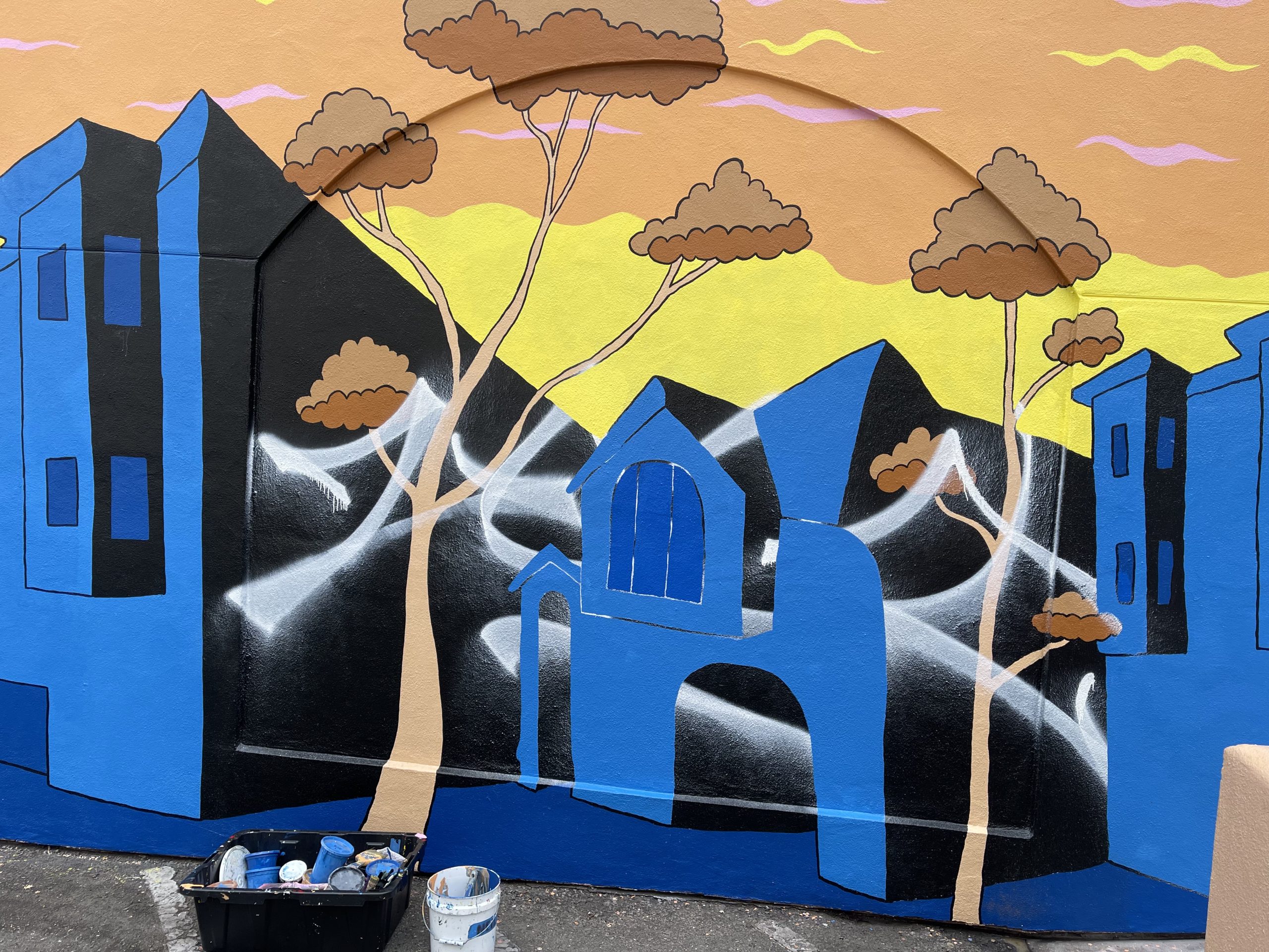 Street mural with blue buildings, trees, and a yellow sky. A paint bucket and supplies sit at the base of the wall.