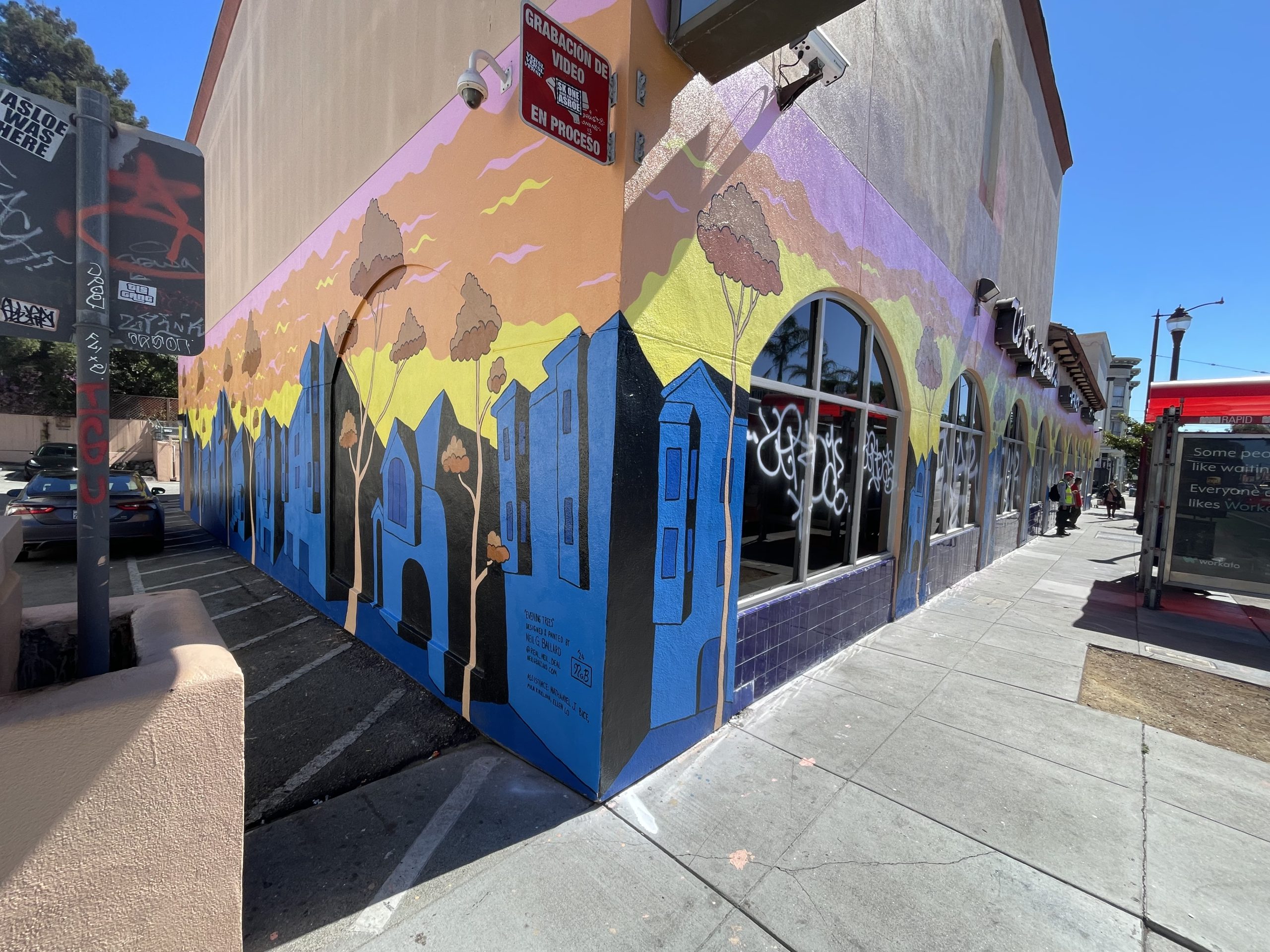 A vibrant mural with abstract buildings and trees decorates the corner of a building, set against a colorful sky above a city sidewalk.