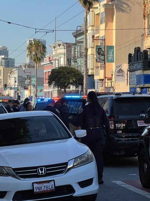 Police officers stand around a vehicle with flashing lights in an urban area during the daytime. People and cars are present in the background.