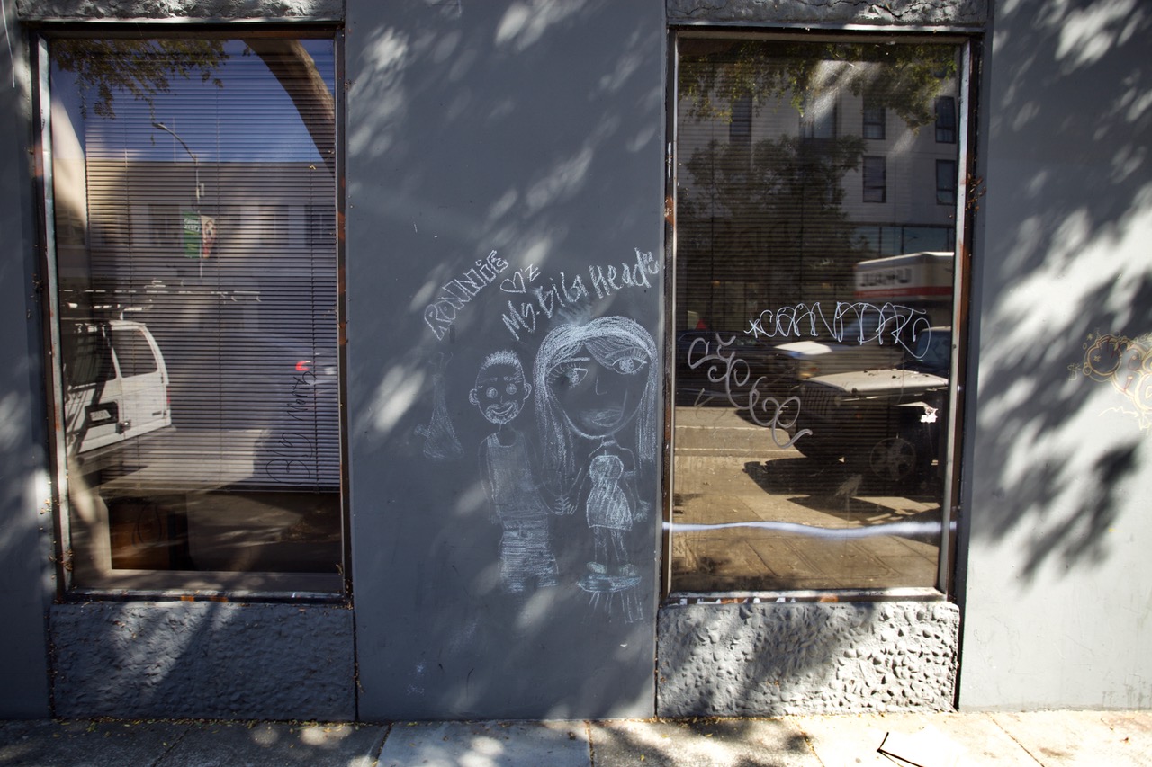 Two large windows flank a chalk drawing on a dark gray wall showing a person with long hair and a child. A van and other buildings are reflected in the windows.