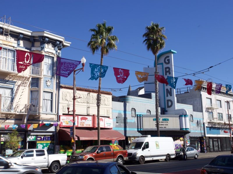 A busy street scene features colorful buildings, a theater with a sign reading "GRAND," parked cars, and vibrant, hanging papel picado.