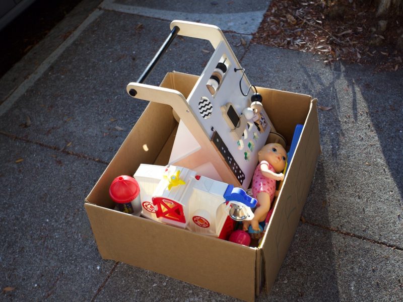 A cardboard box filled with assorted children's toys is placed on a sunlit concrete sidewalk.