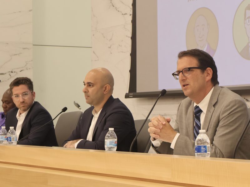 Four District 5 candidates seated at a panel discussion table in a conference room; three men in suits are visible in the foreground.
