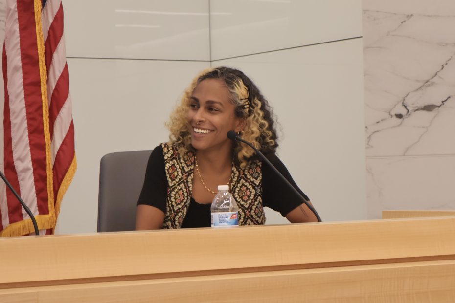 A person with curly hair sits at a desk with a microphone, smiling. An American flag is visible in the background.