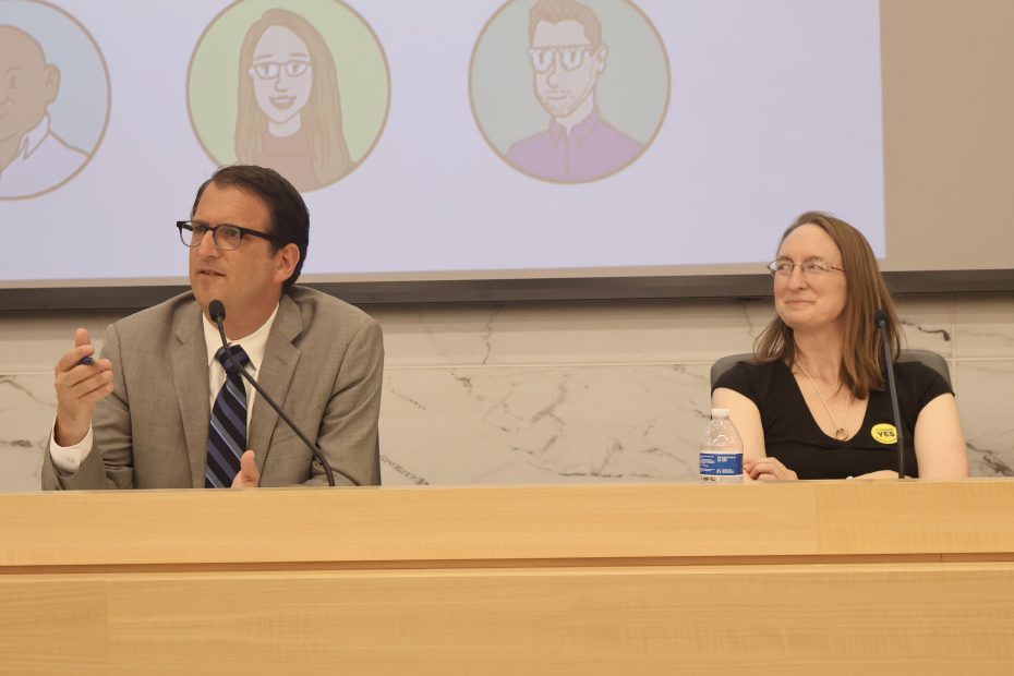 A man in a suit speaks into a microphone at a panel. A woman next to him smiles, seated at a table with a water bottle. A screen in the background shows illustrated avatars.