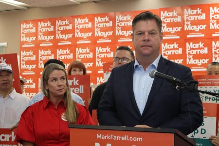 A person stands confidently at a podium speaking at a lively campaign event. Supporters in the background wave "Farrell for Mayor" signs, rallying behind the charismatic candidate.
