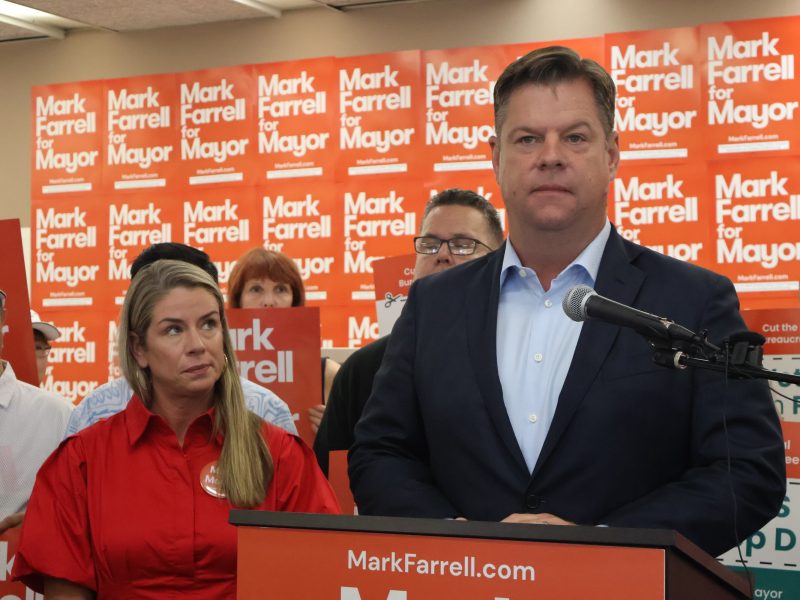 A person stands confidently at a podium speaking at a lively campaign event. Supporters in the background wave "Farrell for Mayor" signs, rallying behind the charismatic candidate.