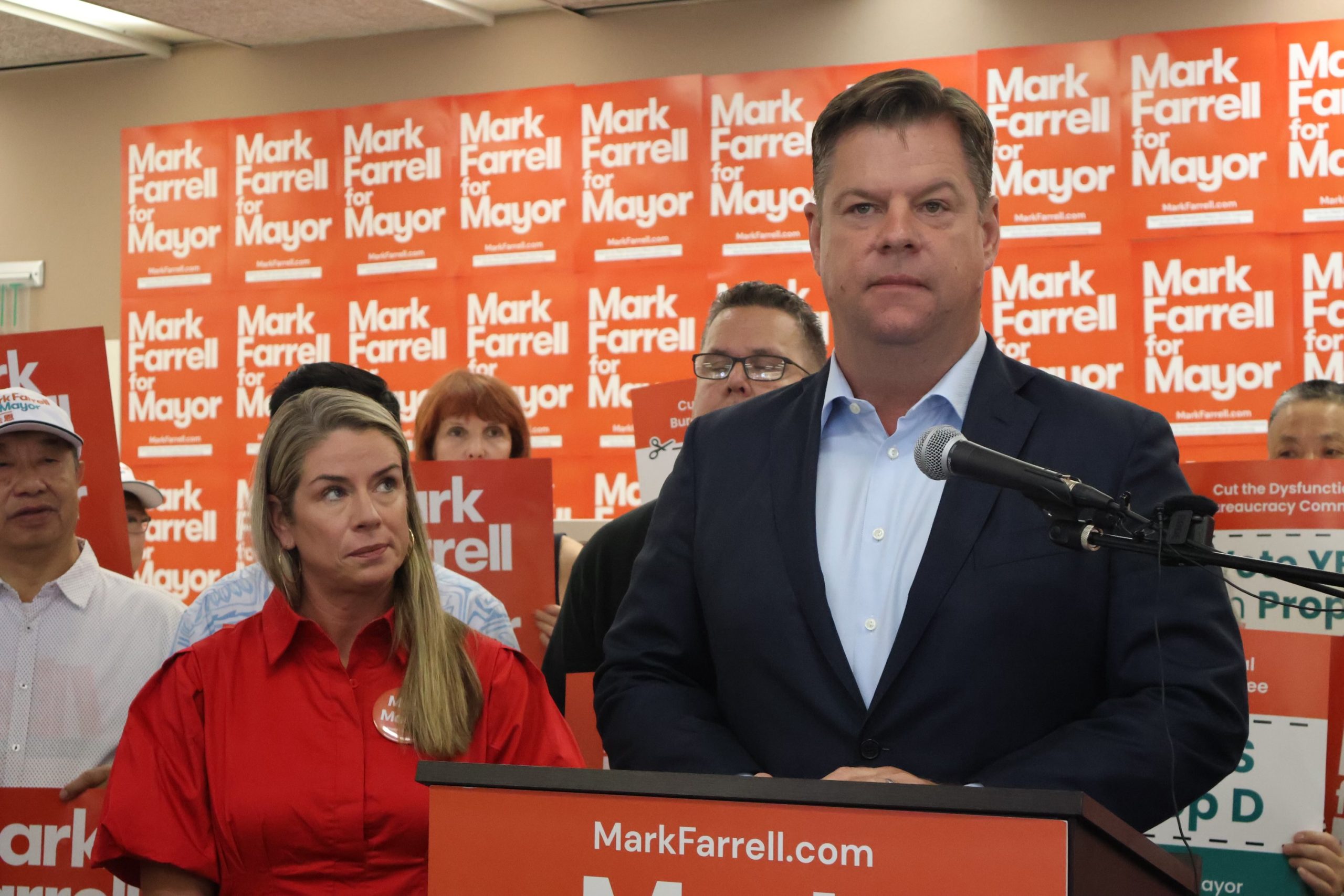 A person stands confidently at a podium speaking at a lively campaign event. Supporters in the background wave "Farrell for Mayor" signs, rallying behind the charismatic candidate.