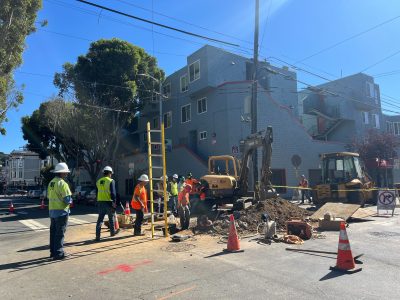 Construction crew and machinery working at a street intersection, with traffic cones and equipment surrounding a utility or maintenance site.