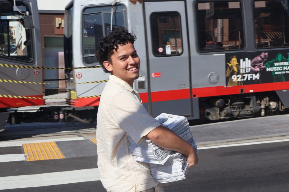 A person smiles while carrying a stack of newspapers, crossing a street with a tram in the background.