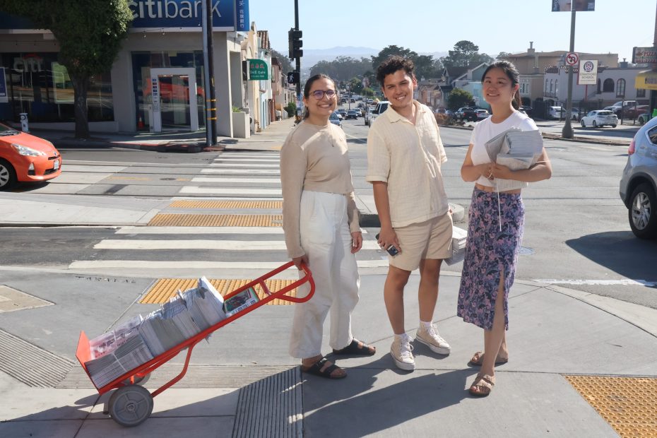 Three people stand at a crosswalk with newspapers on a red trolley.