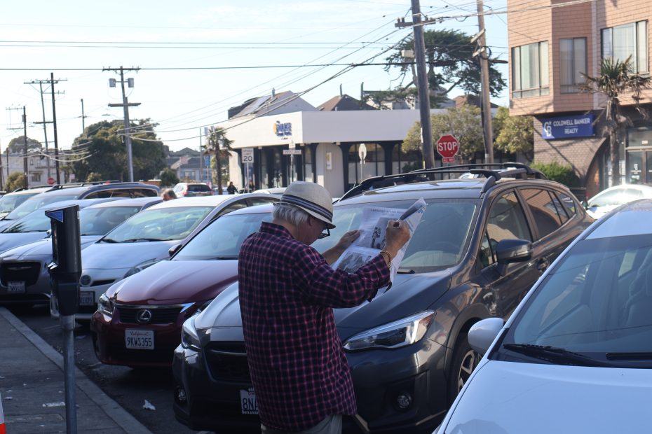 Person in a hat reads a newspaper in a parking lot on a sunny day with parked cars and buildings in the background.