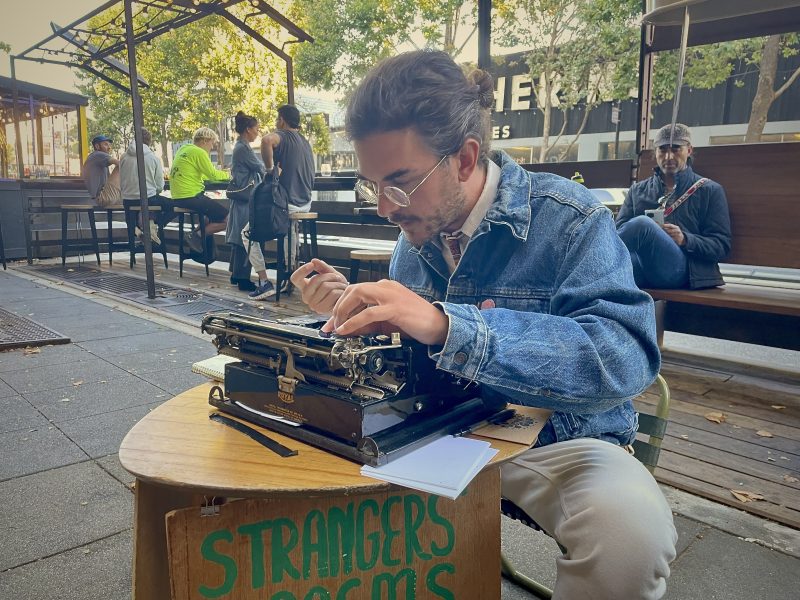 Person typing on a typewriter at an outdoor table with a sign "Stranger's Poems." Others are seated nearby, and trees can be seen in the background.
