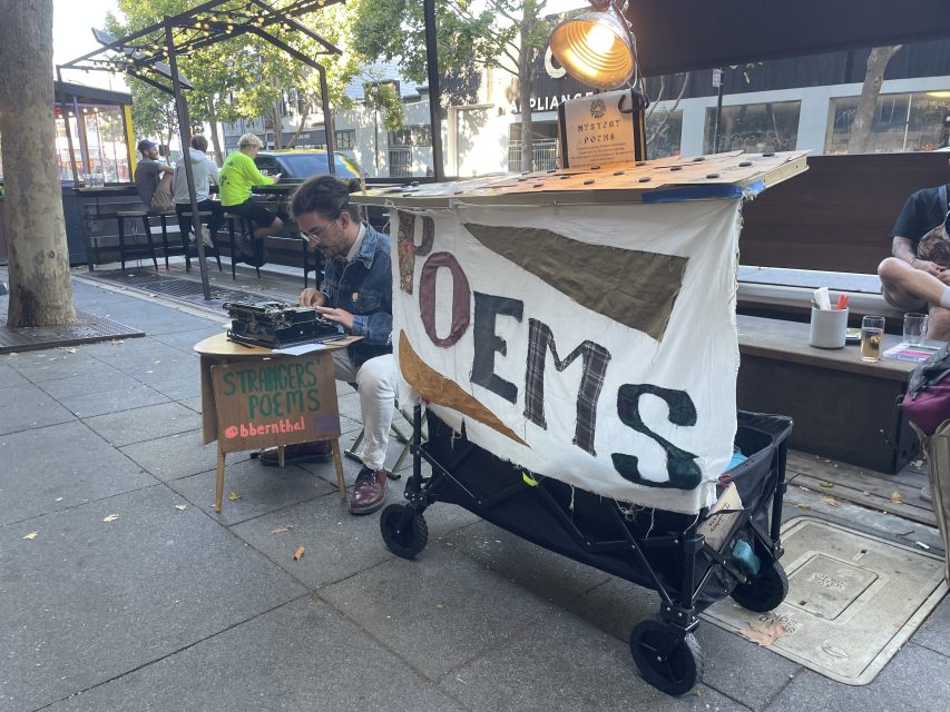 A person types on a typewriter at an outdoor poetry stand with a sign reading "POEMS" on a fabric banner. Nearby is a table with a sign: "Strangers Poems.