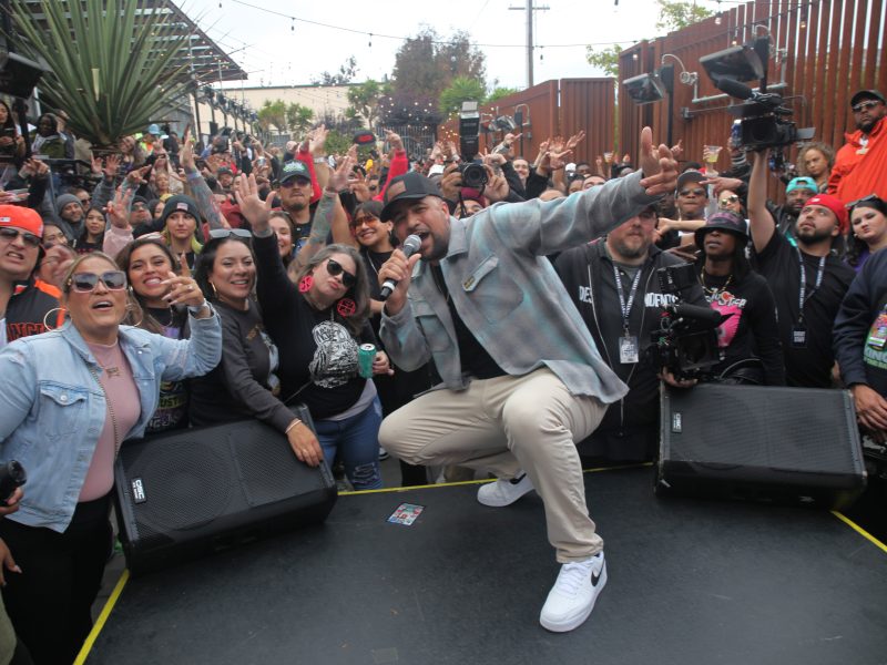 Man holding a microphone kneels on stage while surrounded by a cheering crowd at an outdoor event.