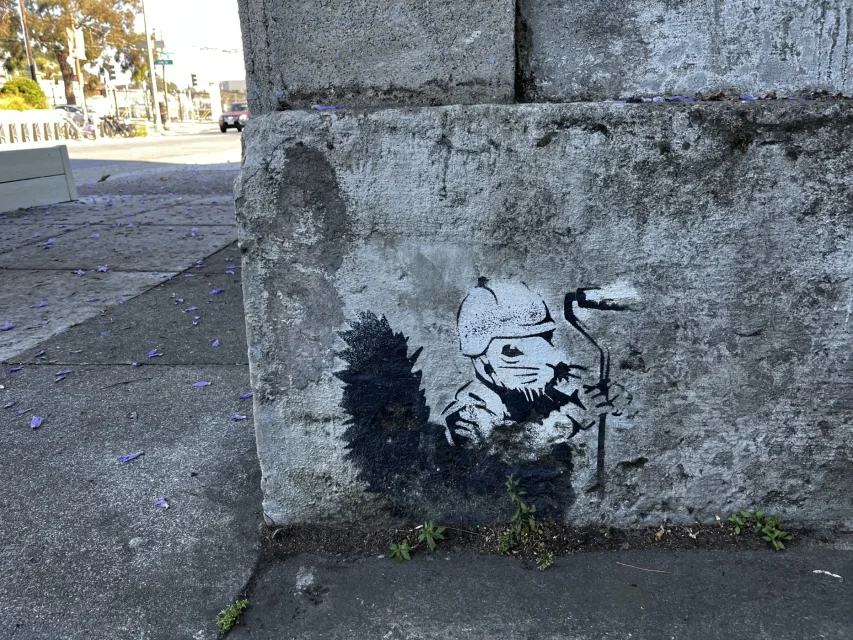 Street art of a soldier holding a paint roller, painted on a concrete wall, with purple flower petals scattered on the ground.