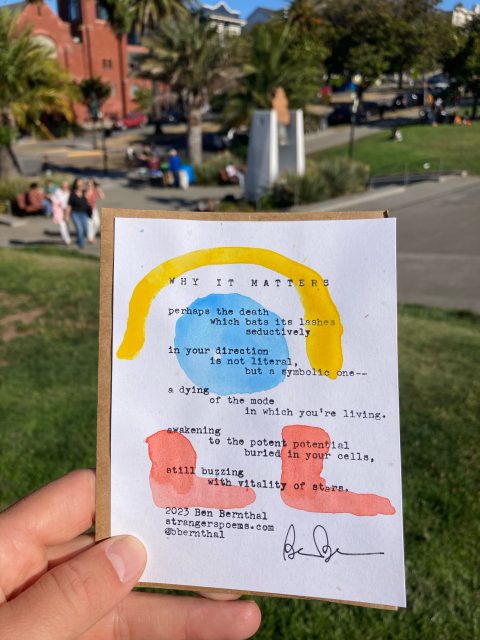 A handwritten poem on colorful painted paper is held up in a park setting with grass, trees, and people in the background.