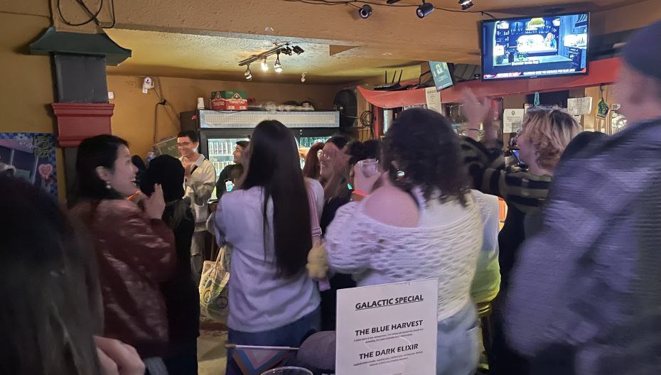 A group of people gathered indoors, some clapping and smiling. A menu with drinks titled "Galactic Special" is visible in the foreground.