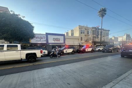 Several police vehicles with flashing lights are parked along a street near a jewelry store on Mission Street.