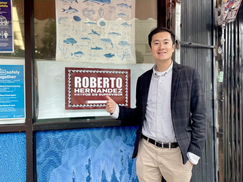 A person stands smiling, holding a sign that reads "Roberto Hernandez for D9 Supervisor" in front of a window with fish illustrations.
