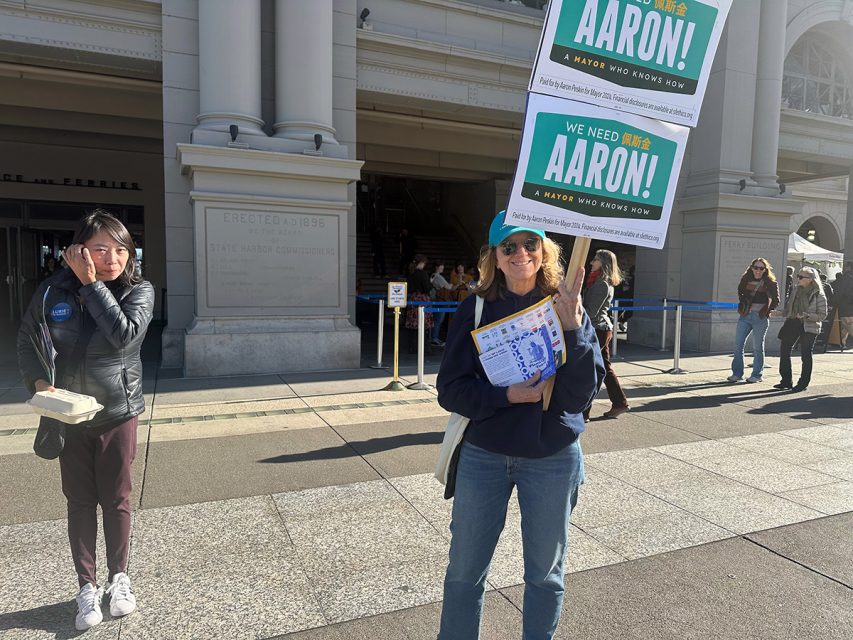 A person holding SF election campaign signs and flyers stands outside a building, while another individual strolls by with a phone and a takeout container.