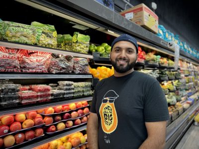 A person stands in front of a grocery store produce section, wearing a black t-shirt and a black cap, surrounded by fruits and vegetables.