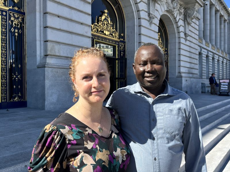 A man and a woman stand together outside a large building with ornate doors and stone steps.